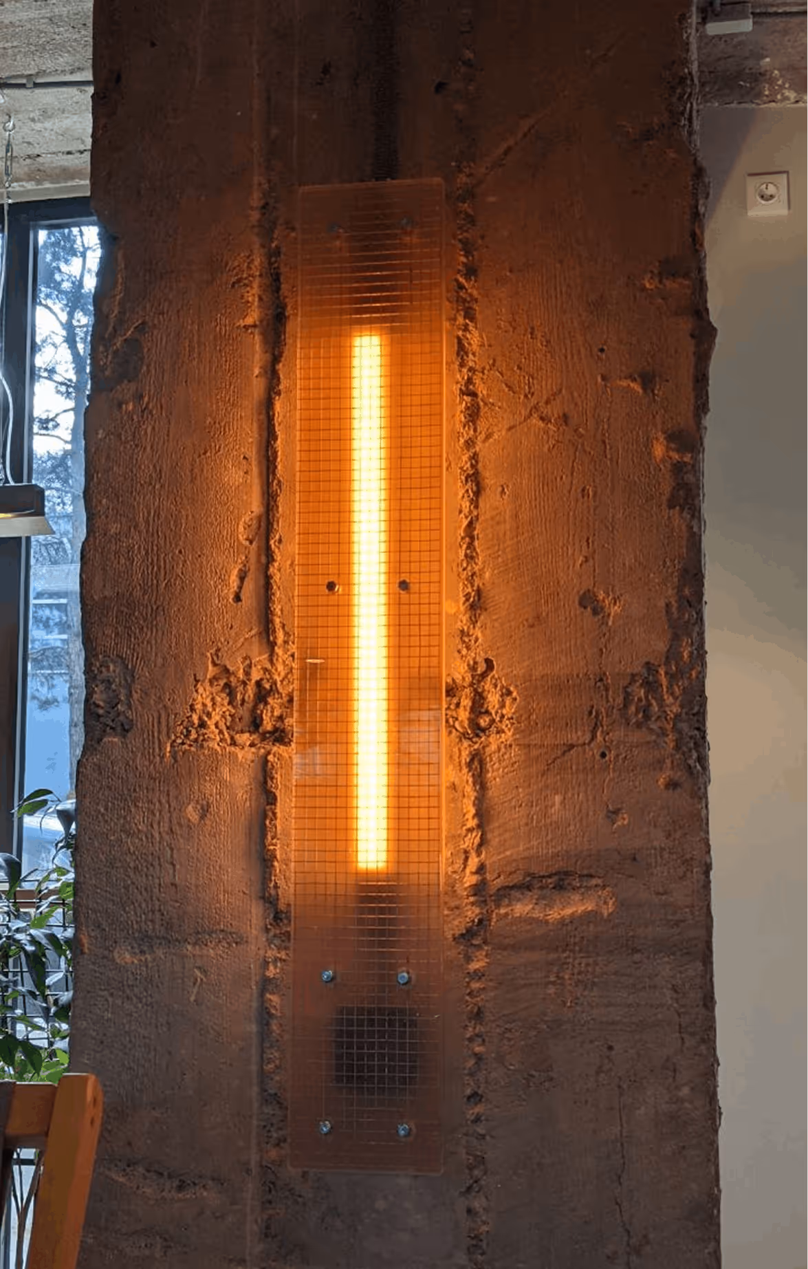 Vertical warm light strip mounted on a rough textured concrete wall near a window with plants.