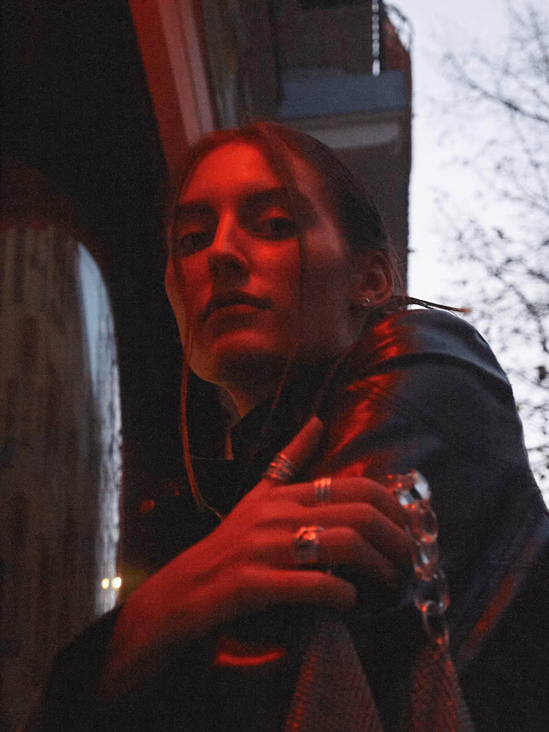 Woman in dark jacket under red light holding a bag with a chain strap outdoors at dusk.