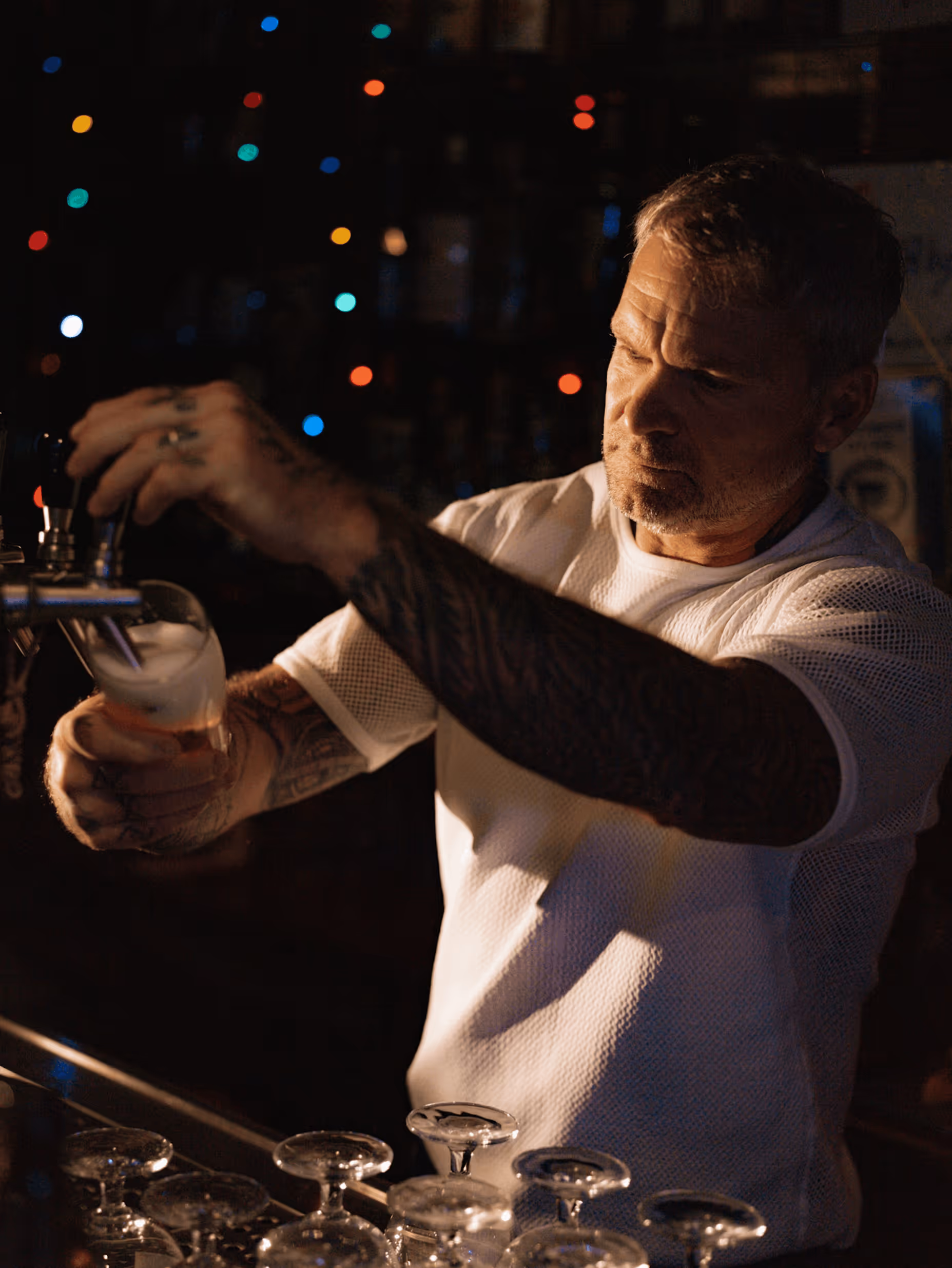 Bartender with tattooed arm pouring beer from tap in dimly lit bar with colorful lights in background.