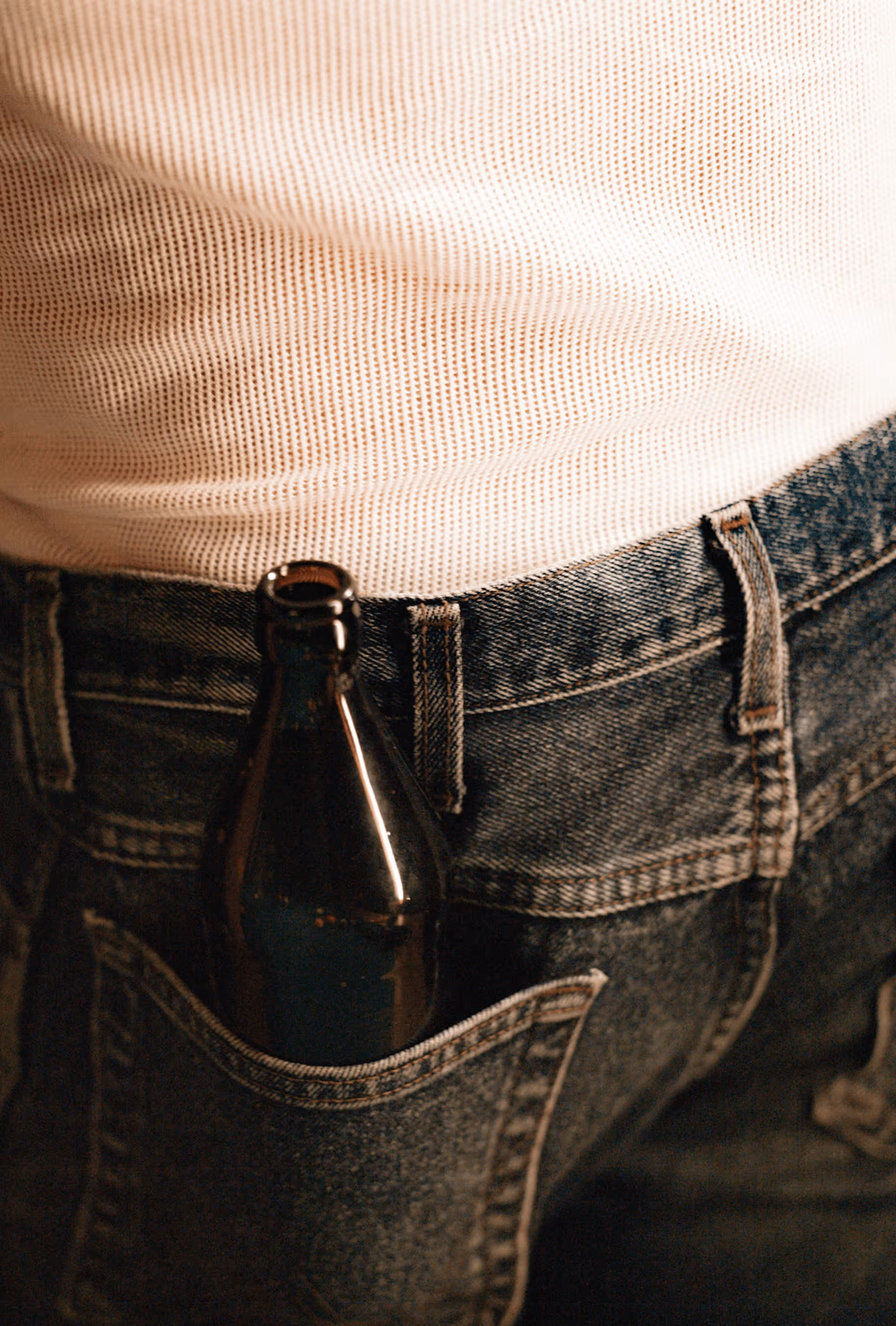 Brown glass bottle tucked into the back pocket of denim jeans worn with a cream shirt.