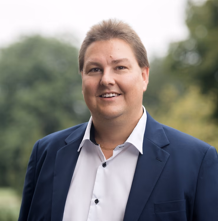 Porträt eines lächelnden Mannes mit kurzem braunem Haar, weißem Hemd und dunkelblauem Sakko vor unscharfem Baumhintergrund.