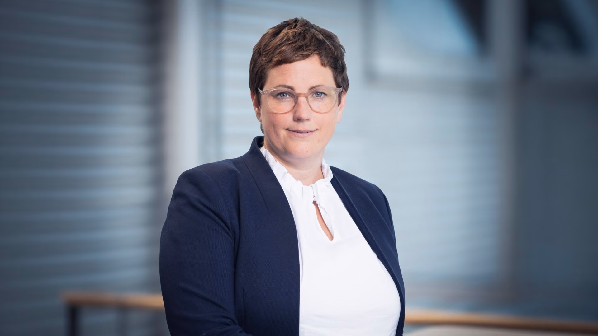 Inga Oehl, die eine weiße Bluse und ein dunkelblaues Sakko trägt, vor einem unscharfen Hintergrund.