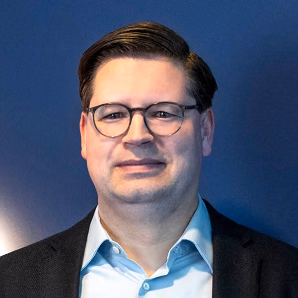 Benjamin Grimm, der ein hellblaues Hemd und dunkle Jacke vor einem blauen Hintergrund trägt.