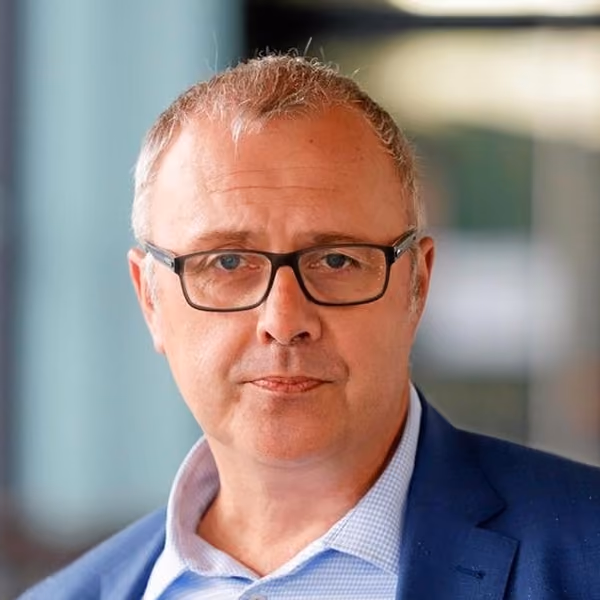 Martin Kaczor, der schwarze Brille trägt und einen blauen Anzug mit hellblauem Hemd anhat, vor unscharfem Hintergrund.