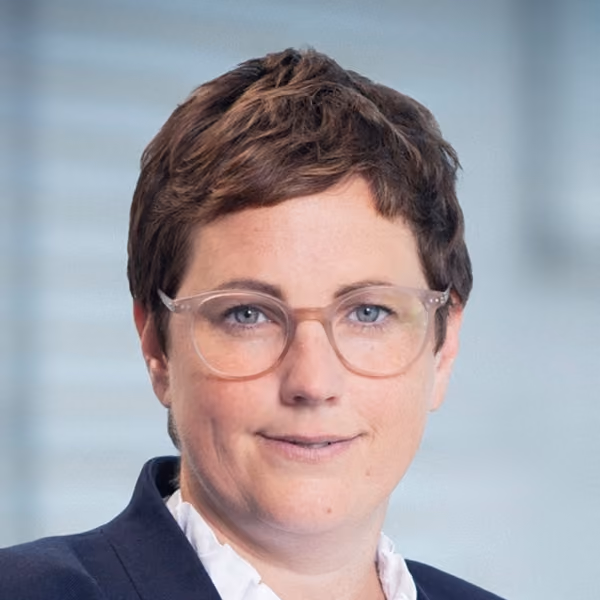 Inga Oehl in dunklem Blazer und weißer Bluse lächelnd in die Kamera blickend, vor unscharfem Hintergrund.