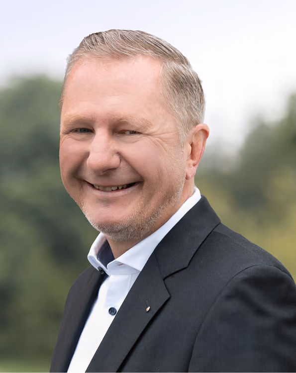Achim Hager, der einen schwarzen Blazer und weißes Hemd trägt, vor einem unscharfen grünen Hintergrund.