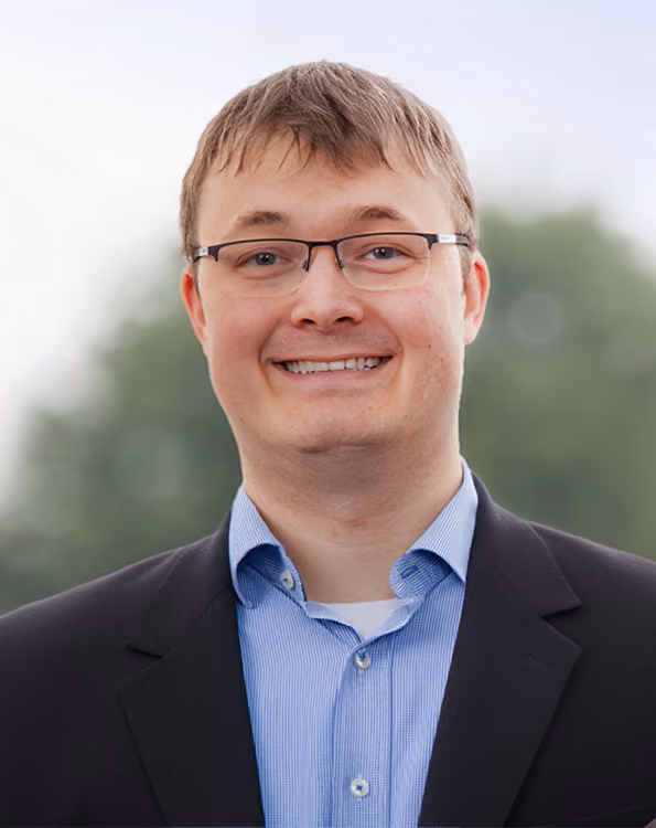 Lorenz Barth, der ein blau gestreiftes Hemd und einen dunklen Blazer trägt, lächelt vor einem unscharfen, grünen Hintergrund.