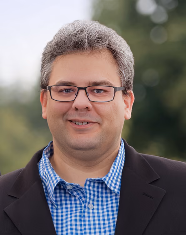 Bernd Büdenbender, der eine Brille, ein blau-weiß kariertes Hemd und ein schwarzes Sakko trägt, vor einem verschwommenen grünen Hintergrund.