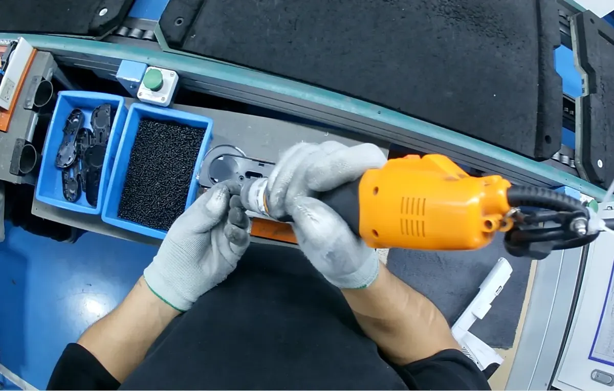 First-person view at an assembly station: gloved hands drive a screw into a black part with an orange electric screwdriver, bins of parts and screws beside them