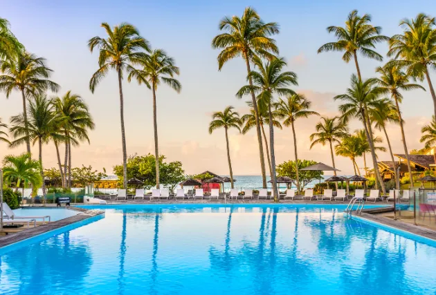 Blick auf den großen Pool des Mahogany Hôtel Résidence & Spa bei Sonnenuntergang, gesäumt von Palmen und Sonnenliegen, mit dem Meer im Hintergrund.