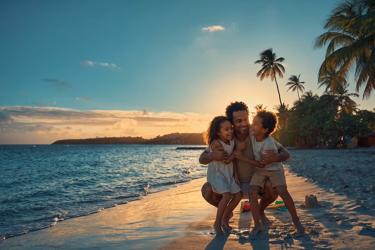 Famille (père, fille, fils) s'étreignant sur la plage au coucher du soleil, avec palmiers, devant Mahogany Hôtel Résidence & Spa, Guadeloupe.