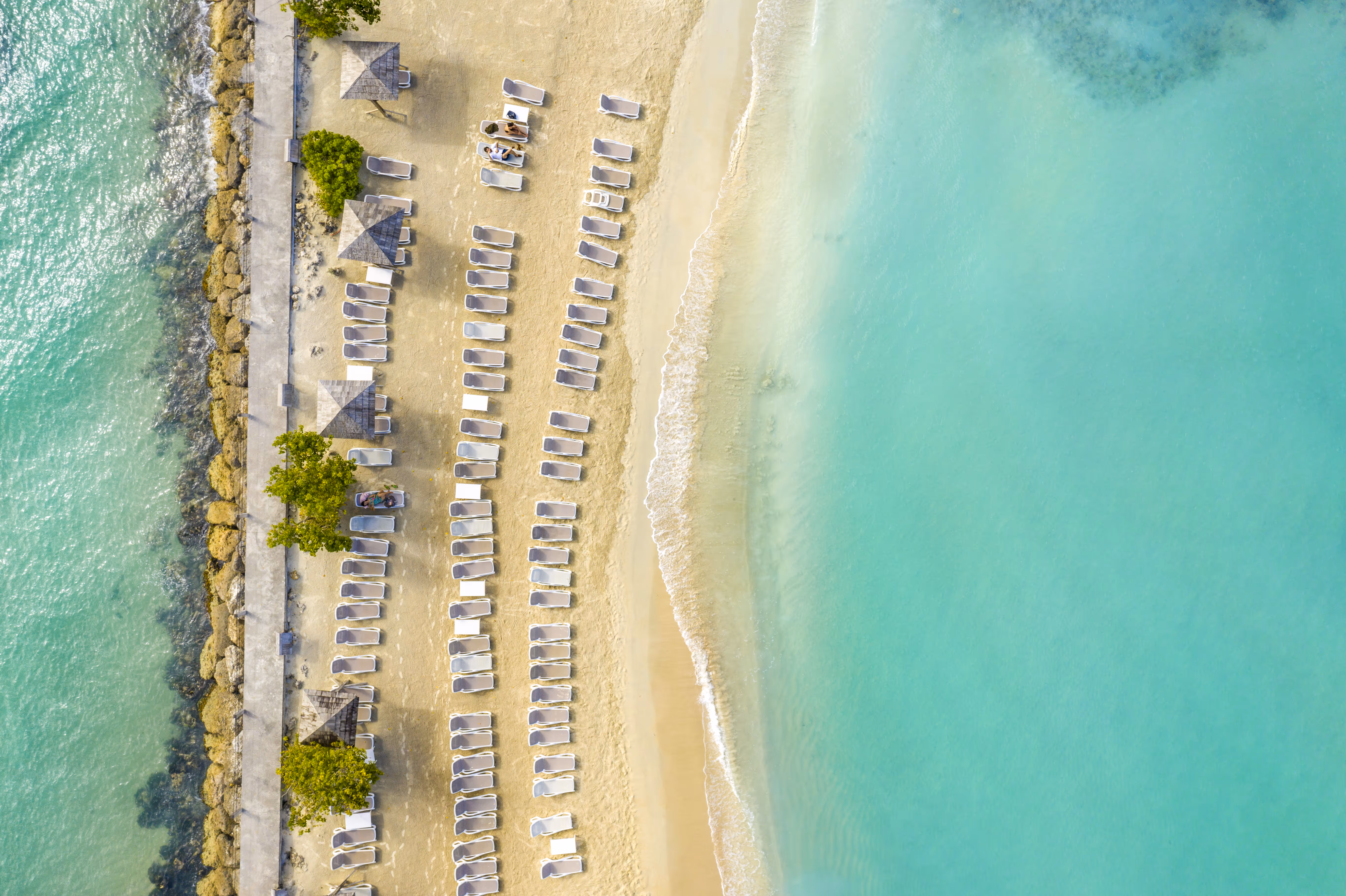 Vue aérienne de la plage de l'hôtel montrant des rangées de chaises longues et la jetée rocheuse. Mahogany Hôtel Résidence & Spa, Le Gosier, Guadeloupe.