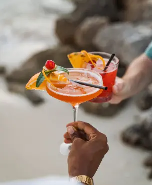 Deux mains portant un cocktail tropical rouge et un verre d'eau au bord d'une piscine au Mahogany Hôtel Résidence & Spa, Le Gosier, Guadeloupe.