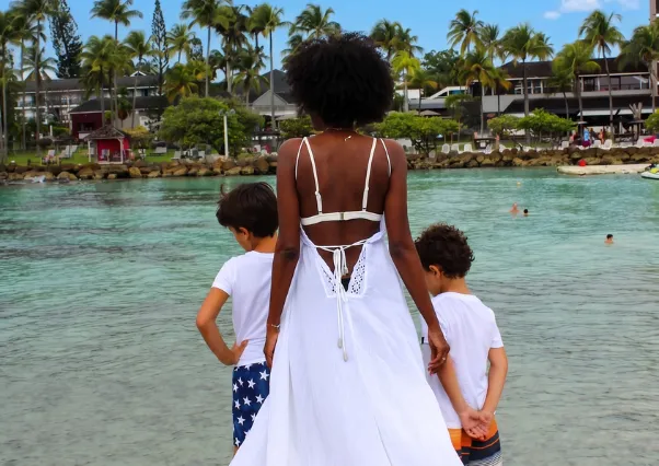 Eine Frau in einem weißen Kleid und zwei Jungen blicken an der Pointe de la Verdure vor der Anlage des Mahogany Hôtel Résidence & Spa auf das Meer.