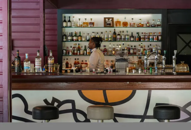 Intérieur d'un bar avec serveur de dos, étagères d'alcools et tabourets, Mahogany Hôtel Résidence & Spa, Le Gosier.