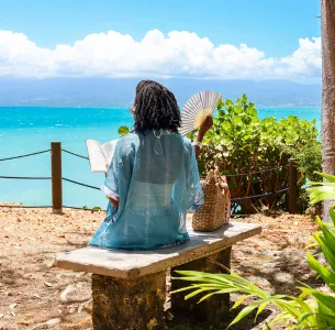 Rückenansicht einer Gästin, die auf einer Steinbank sitzt, ein Buch liest und einen Fächer hält, mit Blick auf das türkisfarbene Meer im Mahogany Hôtel Résidence & Spa.