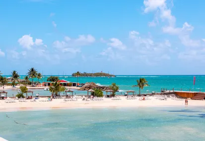 Panorama der Bucht des Mahogany Hôtel Résidence & Spa mit dem feinen Sandstrand, dem Bootssteg und der vorgelagerten Insel unter sonnigem Himmel.