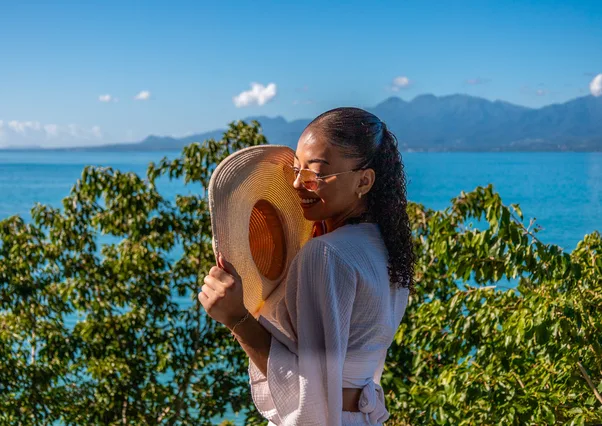 Femme souriante tenant un chapeau de paille devant la mer turquoise et les montagnes, Le Gosier, Guadeloupe.