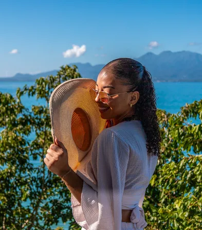 Femme souriante tenant un chapeau de paille devant la mer turquoise et les montagnes, Le Gosier, Guadeloupe.