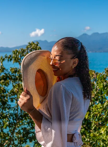 Femme souriante tenant un chapeau de paille devant la mer turquoise et les montagnes, Le Gosier, Guadeloupe.