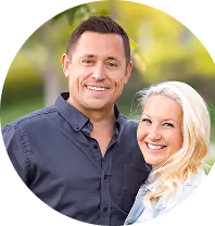 Smiling man in dark shirt and woman with blond hair posing outdoors with blurred green background.