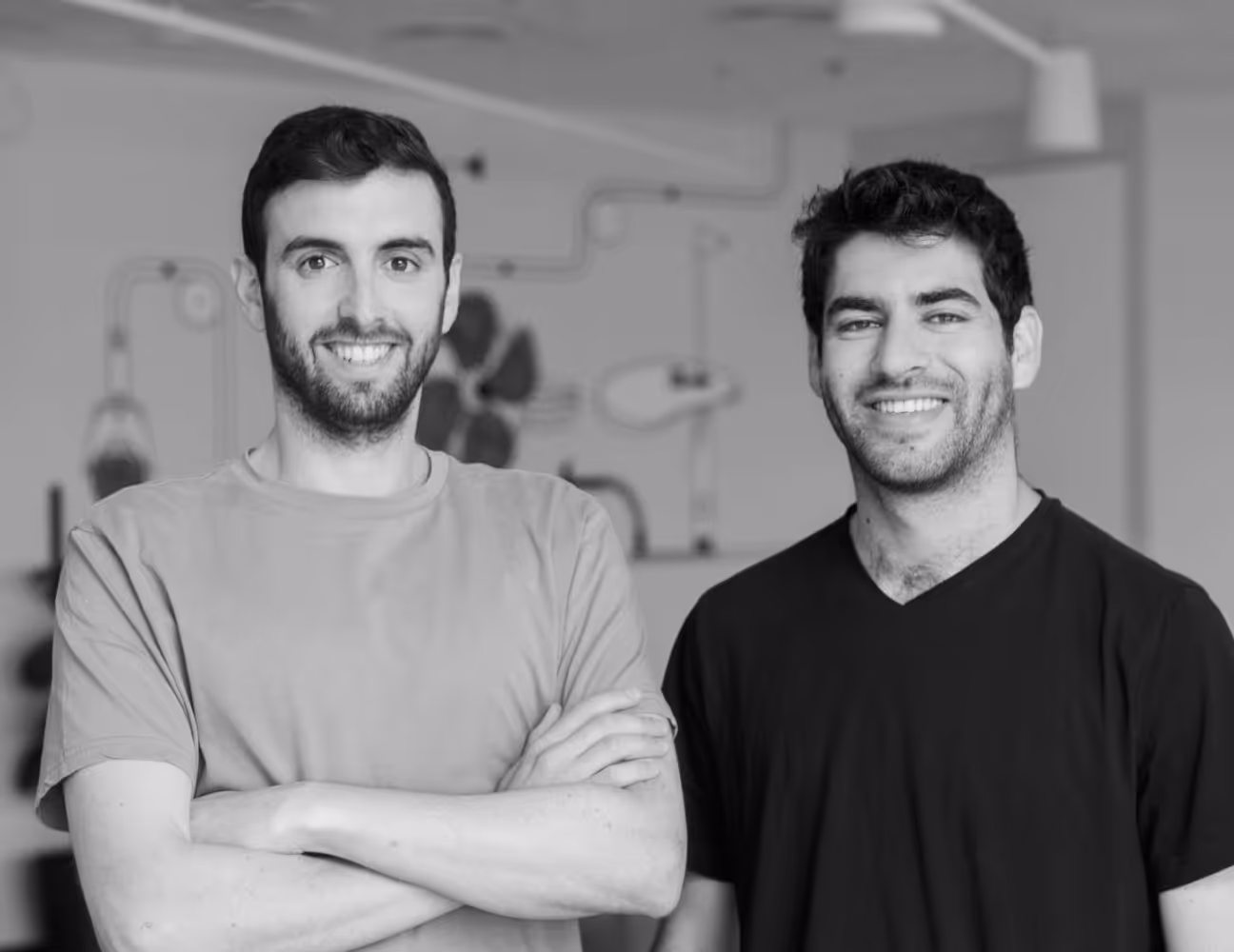 Two young men posing and smiling indoors, one with arms crossed wearing a light shirt, the other in a dark shirt.