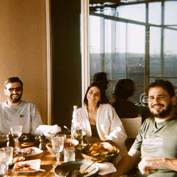 Three people smiling and sitting at a table with food and drinks in a sunlit room.