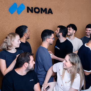 Group of young people socializing and smiling under a NOMA logo on a brown wall.