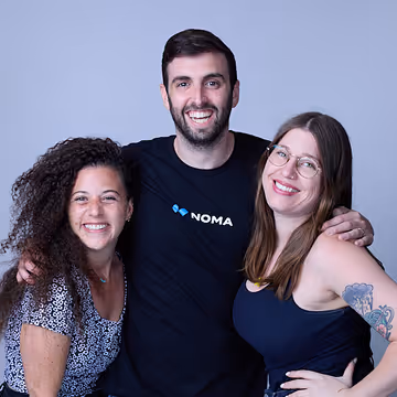 Smiling man in NOMA shirt standing with two cheerful women against a light gray background.