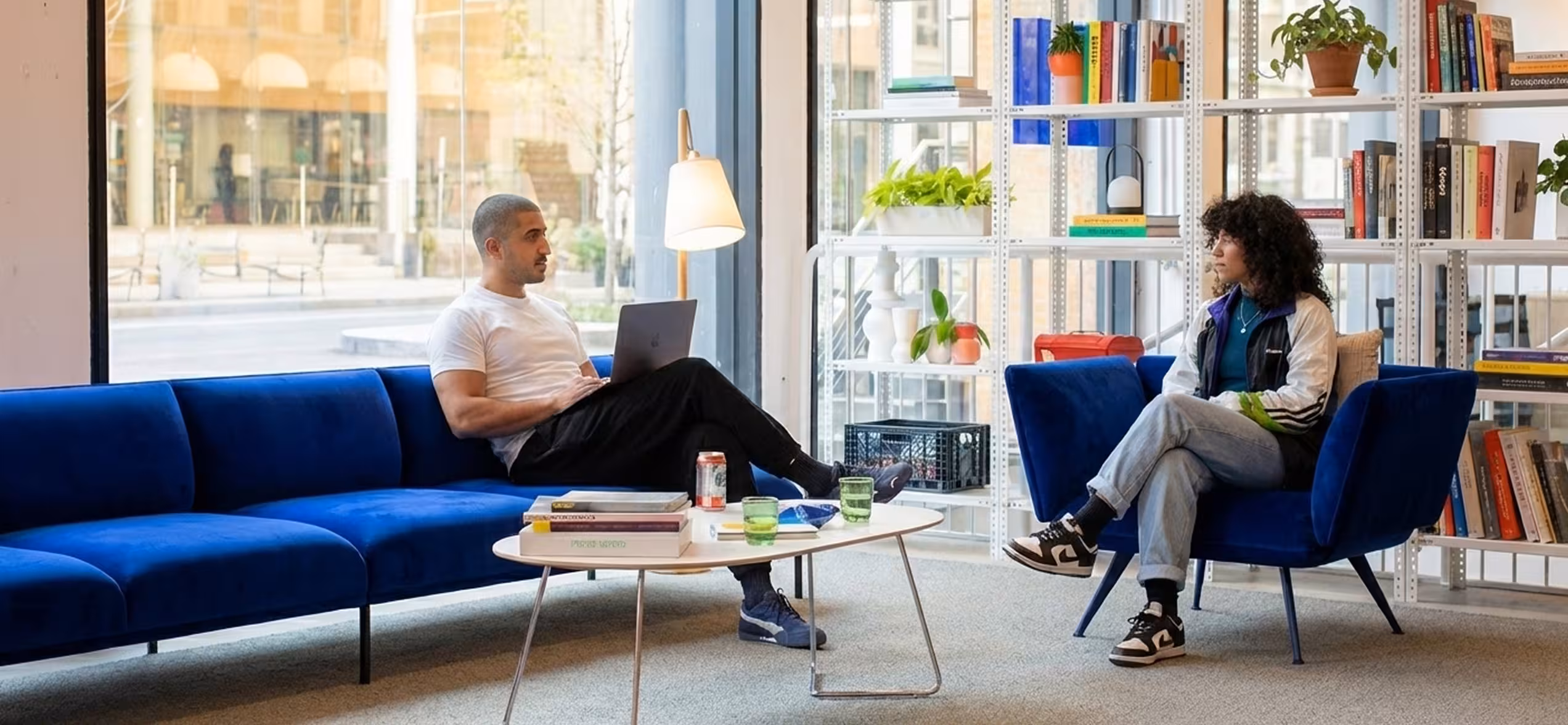 Two people sitting on blue chairs in a modern room with bookshelves and large windows.