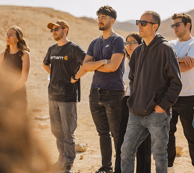Six adults standing outdoors on dry terrain, attentively looking at something off-frame.