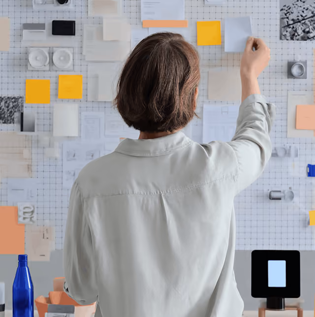 Person organizing various sticky notes and papers on a grid wall viewed from behind.