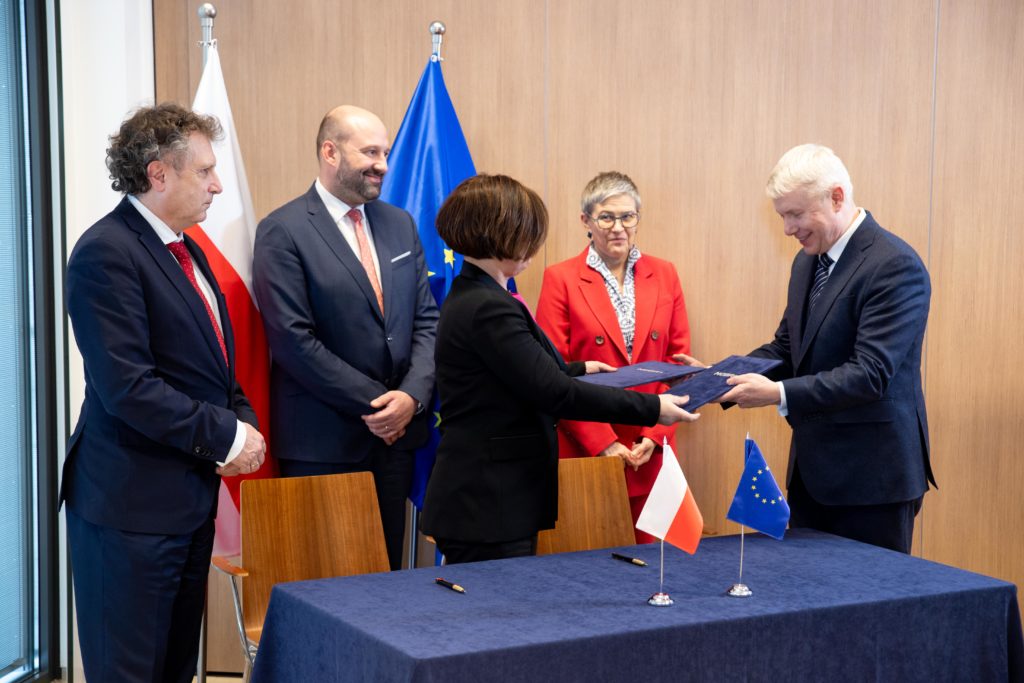 Two signatories exchange folders across a table with Polish and EU flags as three officials look on