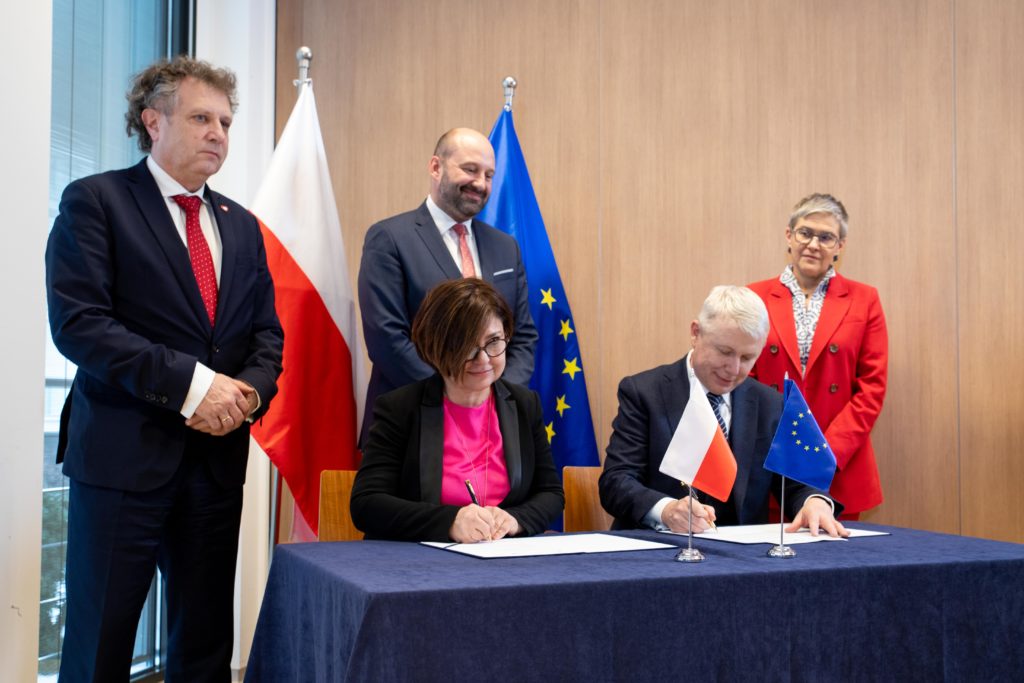 Two representatives sign the STEP project agreement at a table with Polish and EU flags, three officials standing behind