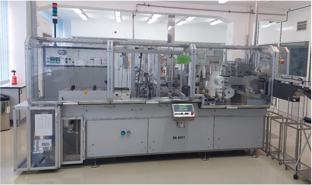 CAM blister packaging machine with control panel and film reels behind clear guarding in a production room
