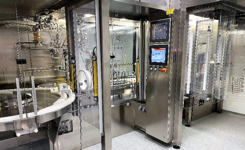 Stainless steel vial filling and capping machine with touchscreen controls and rotary conveyor behind glass panels
