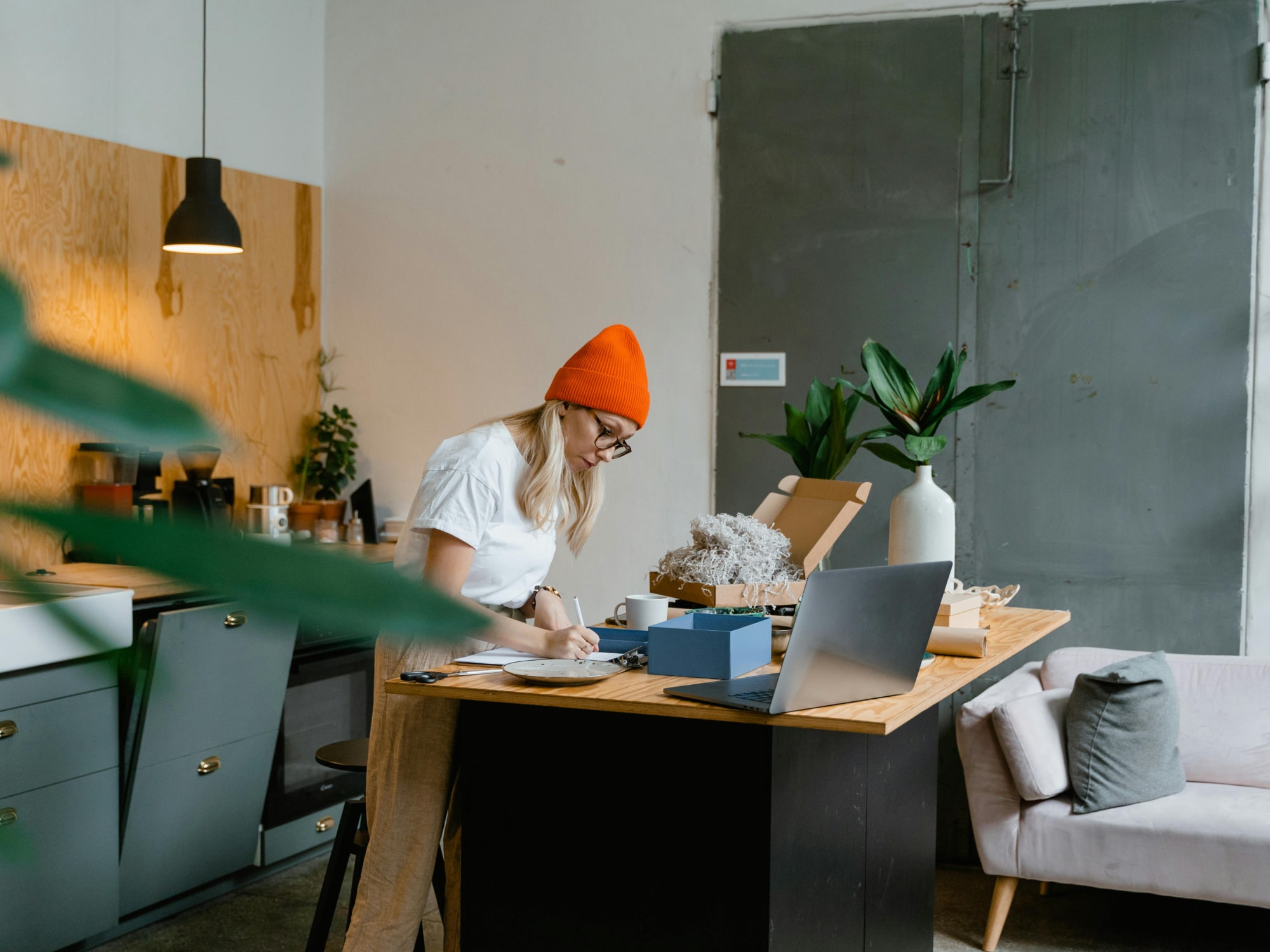Woman in orange beanie writing at a wooden table with laptop, plants, and open box in modern kitchen.