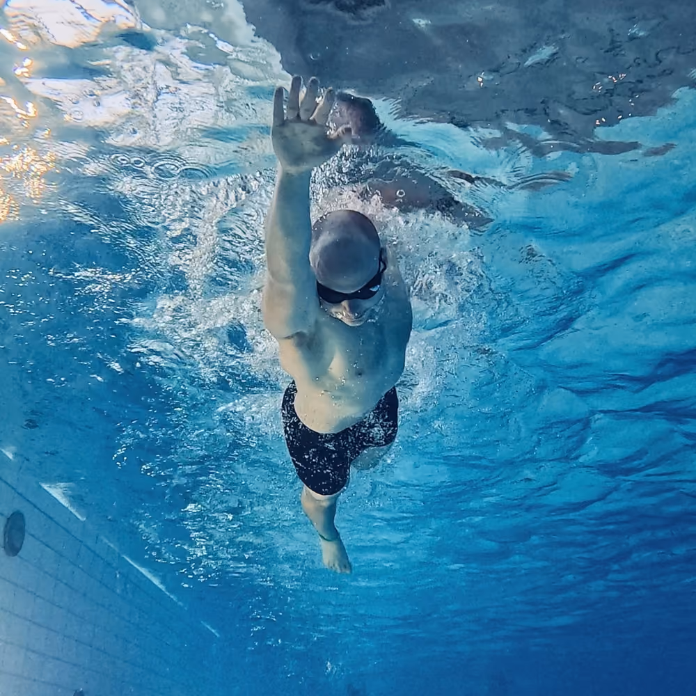 Ein Schwimmer unter Wasser in einem Pool, der mit einer Hand nach vorne schwimmt und eine  Schwimmbrille trägt.
