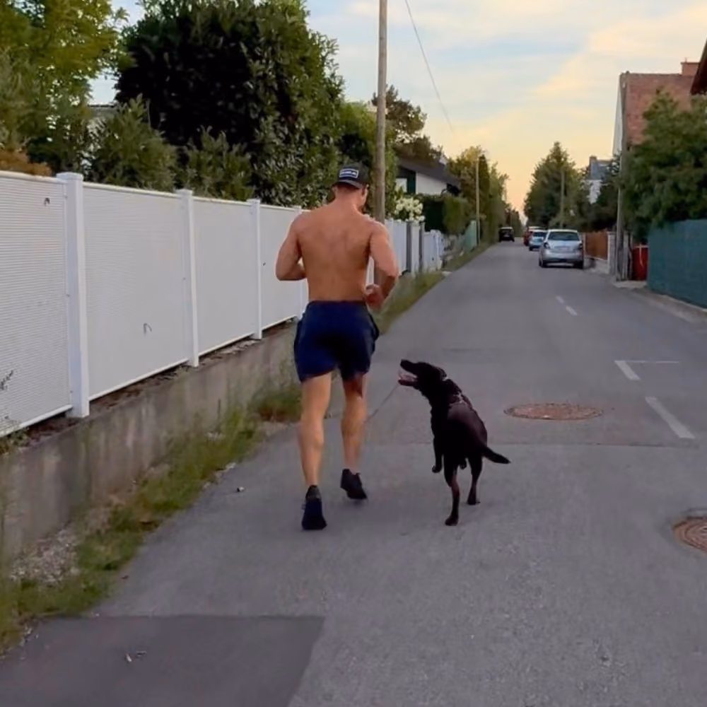Ein muskulöser Mann joggt auf einer ruhigen Wohnstraße, begleitet von einem Hund, der neben ihm läuft und zu ihm aufblickt.