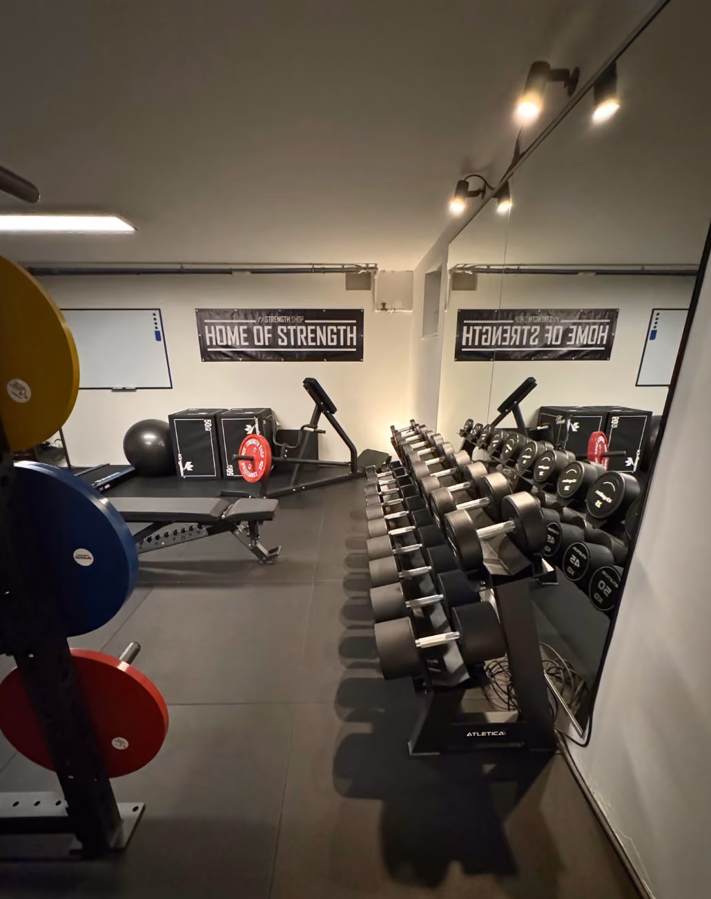 Innenansicht eines Fitnessraums mit Kurzhantelregal vor einem großen Spiegel, Gewichtsscheiben auf einer Halterung links und Trainingsbank im Hintergrund, an der Wand hängt ein Schild mit der Aufschrift 'HOME OF STRENGTH'.