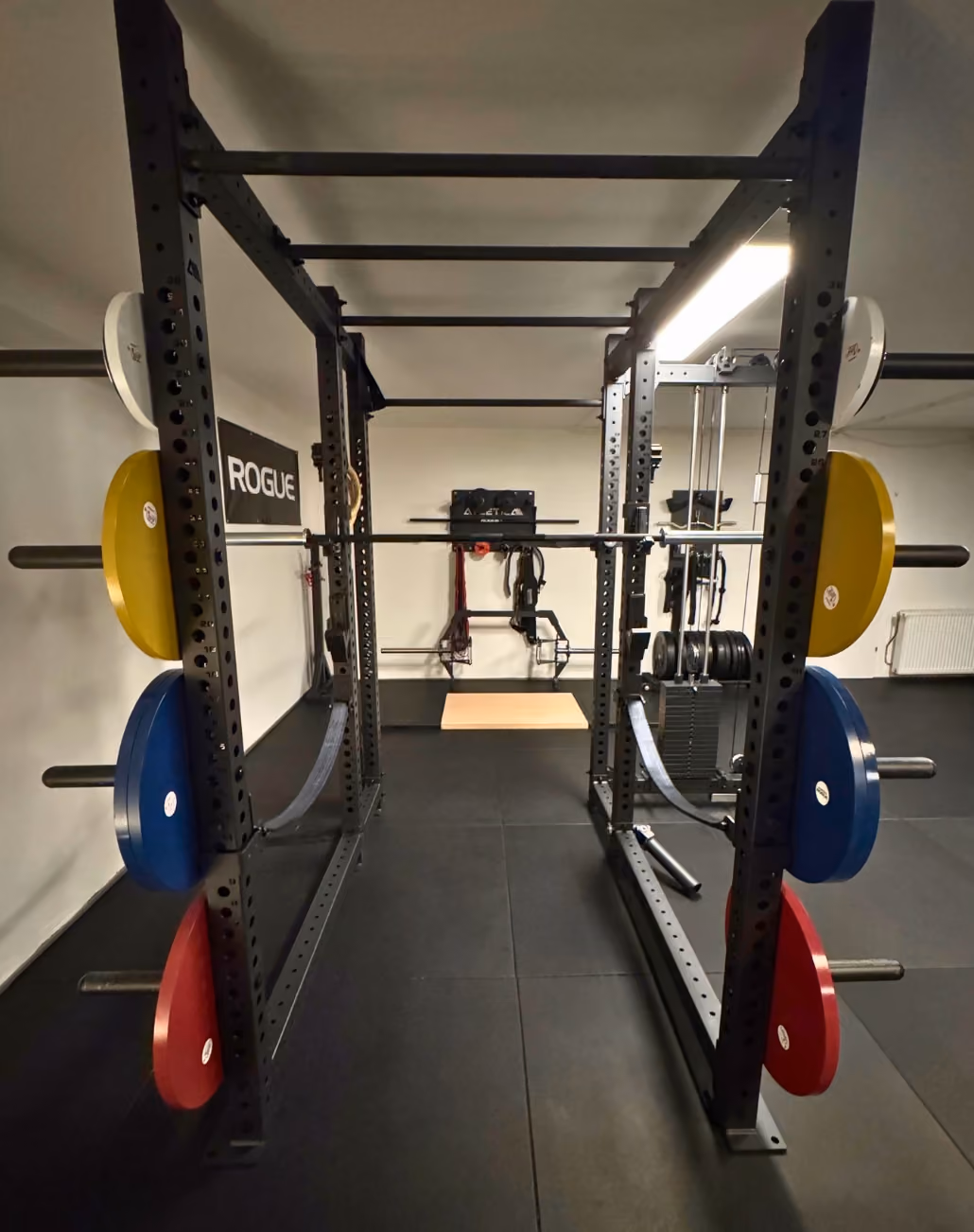 Schwarzer Kraftturm im Fitnessstudio mit verschiedenen Gewichtsscheiben in Weiß, Gelb, Blau und Rot an den Seiten, einer Langhantelstange und Trainingsausrüstung im Hintergrund.
