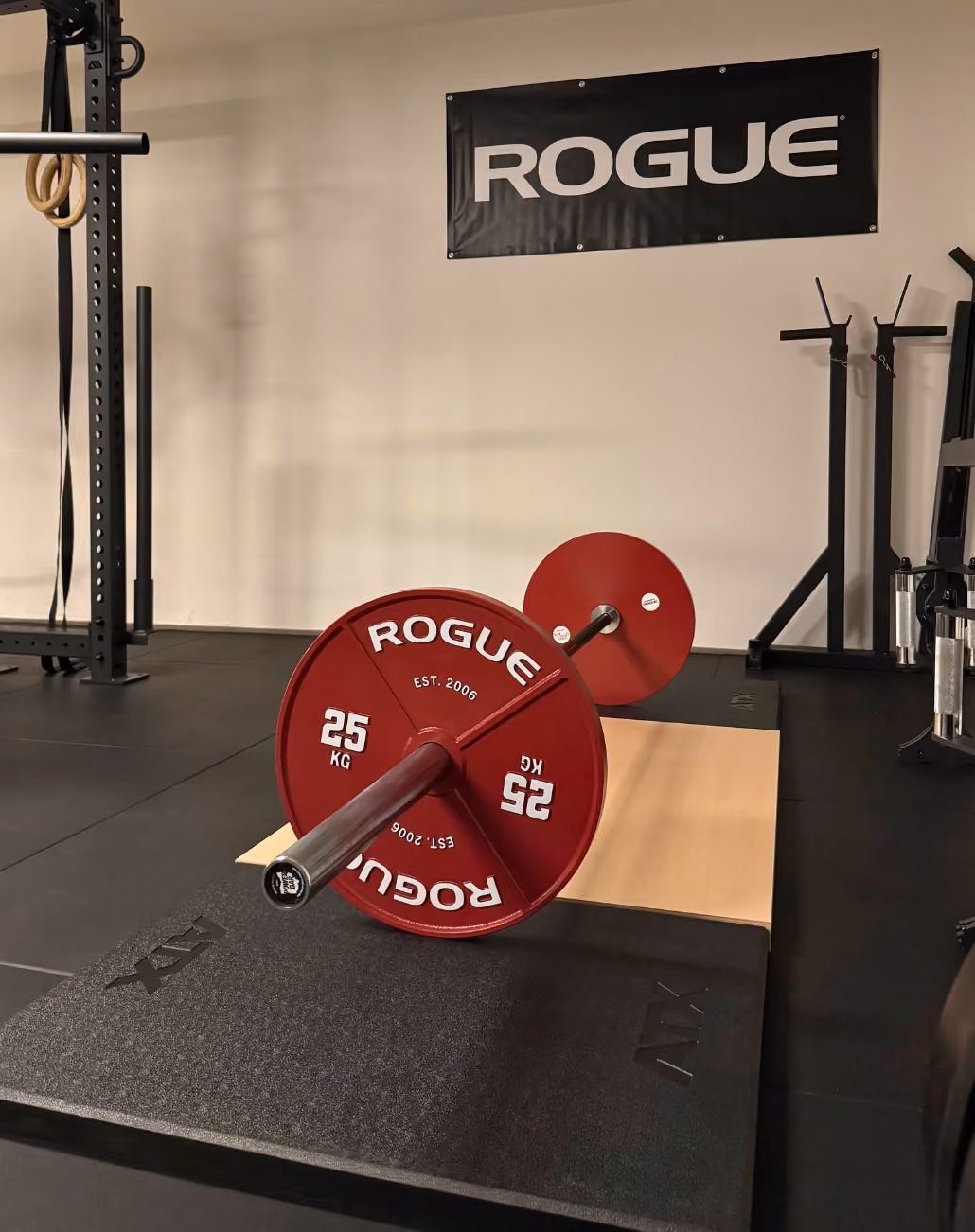 Langhantel mit roten Rogue Gewichtsscheiben zu je 25 kg liegt auf einem schwarzen und einem hellbraunen Bodenmattenbereich in einem Fitnessstudio.