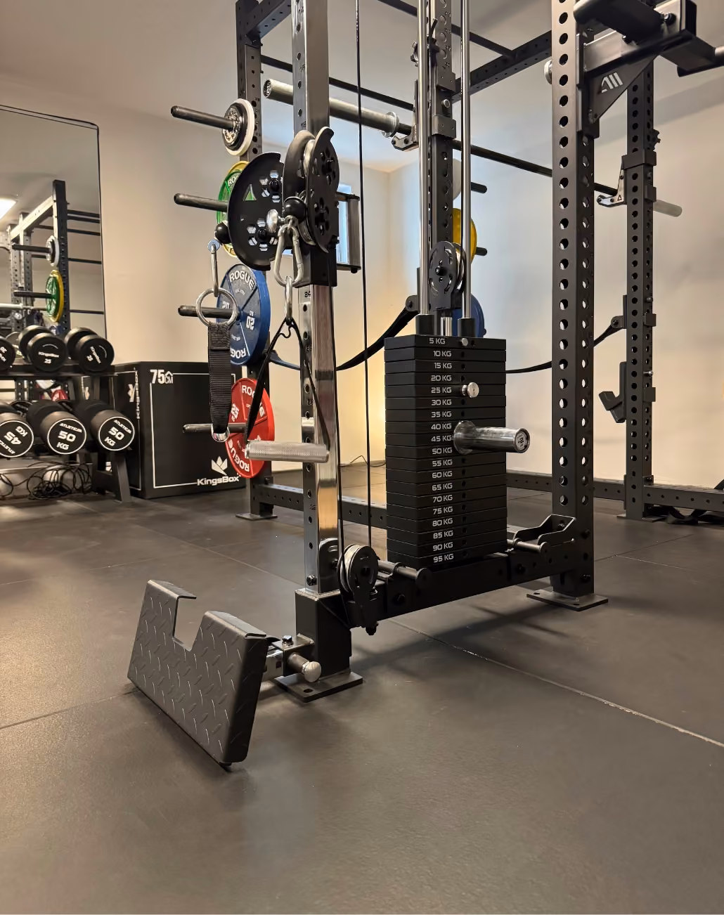 Fitnessstudio mit Hanteln, einer Langhantel mit farbigen Gewichtsscheiben, einem Gewichtsturm mit aufsteigenden Gewichtsplatten von 5 bis 95 Kilogramm und einer Kabelzugmaschine auf grauem Gummiboden.
