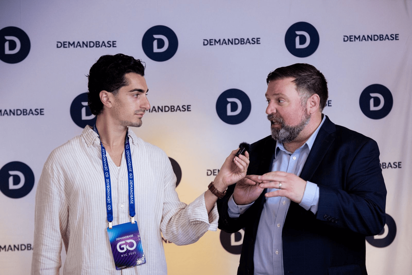 Man with lanyard interviews another man in a suit in front of a Demandbase backdrop.