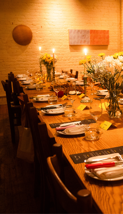 Long wooden table set for dinner with flowers, candles, plates, utensils, and glasses in warm lighting.