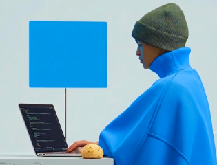 Person in blue coat and green beanie working on a laptop at a white table with a small bread roll.