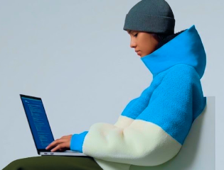 Person in blue and white jacket with gray beanie typing on laptop while sitting on chair.
