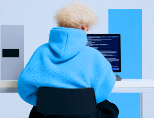 Person in a blue hoodie sitting at a desk, coding on a laptop against a blue background.