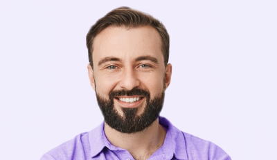 Smiling bearded man with short brown hair wearing a purple shirt against a light background.