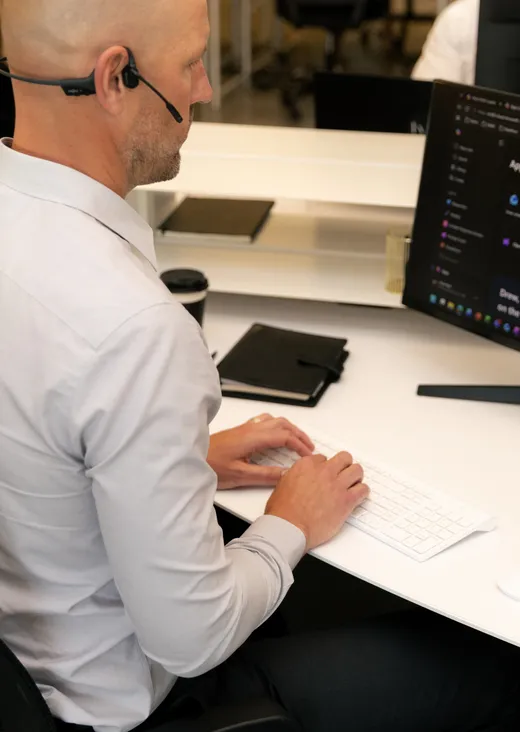 Man wearing a headset typing on a keyboard at a desk with a computer monitor and notebook.